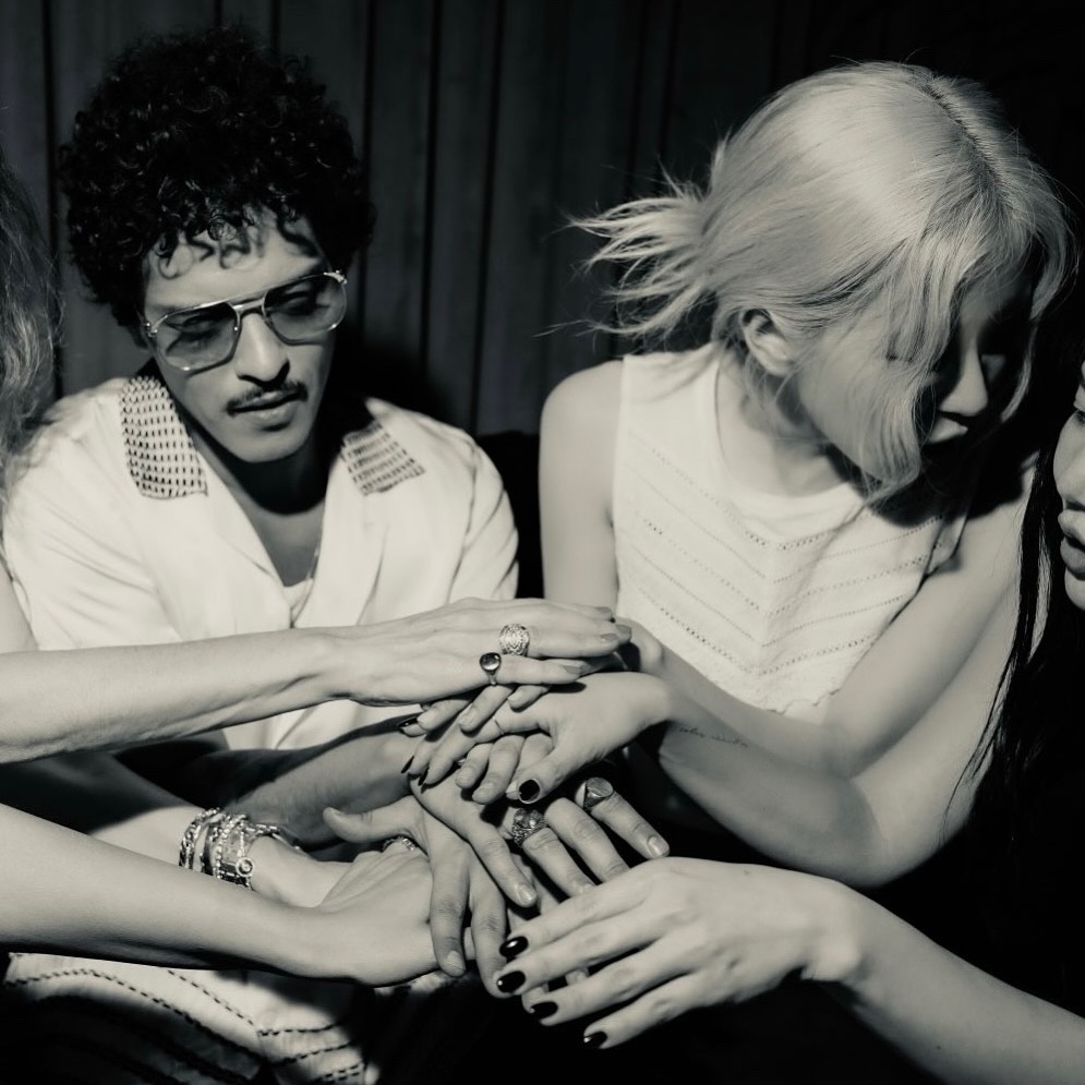 the black and white photograph depicts a group of people, primarily women, with a man centrally positioned.  their hands are layered on top of each other in a gesture of unity or togetherness.  they appear to be wearing rings and other jewelry. the style is reminiscent of 70s or 80s fashion, with the man sporting curly hair and sunglasses.  the overall tone is moody and atmospheric, with a focus on the hands and the interplay of light and shadow. the background is dark and indistinct, drawing attention to the subjects.