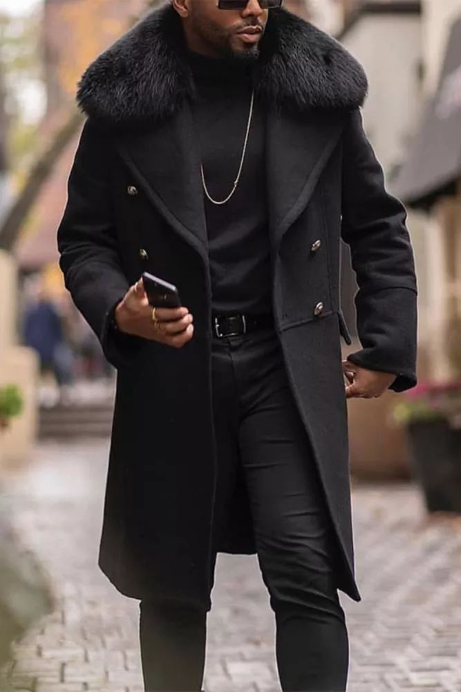 a man is walking down a city street. he is wearing a black coat with a fur collar, sunglasses, and black pants. he is looking down at his phone.