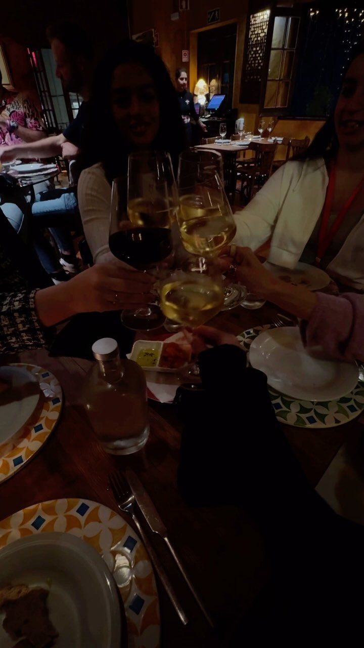 the image shows a group of people toasting with wine glasses at a table in a restaurant. there are plates, silverware, and a liquor bottle on the table. the lighting is dim, creating a warm and intimate atmosphere.