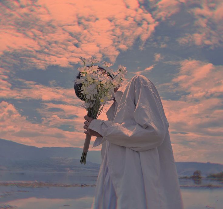 a young man, slightly turned, obscures his face with a bouquet of white daisies. he is dressed in flowing white clothing and stands against a backdrop of a vast, serene landscape featuring a pastel sunset, clouds and distant mountains. the image evokes feelings of peace and tranquility.