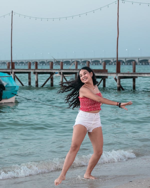 a young woman with long dark hair is joyfully running along a sandy beach near the water's edge. she's wearing a red polkadot crop top and white shorts.  the ocean waves gently lap at the shore. in the background, there's a wooden pier extending over the water, with a boat visible in the distance and a bridge further back. the overall mood is happy, carefree, and summery, suggesting a vacation or getaway. the lighting appears to be either late afternoon or early evening.