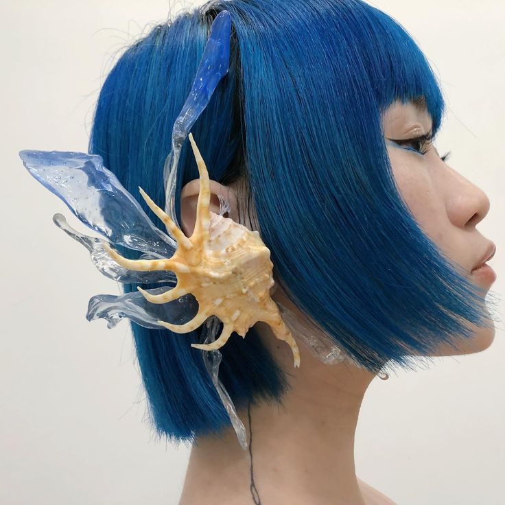 a woman with bright blue hair,  wearing a seashell and clear plastic ear cuff.  she is looking to the right of the frame with a stoic expression.