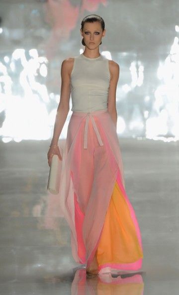 a woman model walks the runway in a white tank top and a flowing pink and orange skirt. she is wearing a pair of lightcolored heels. she is holding a white clutch. she is posed with her arms by her side, looking straight ahead. her hair is slicked back and she is wearing light makeup. the background is a blurry studio shot of a white backdrop.
