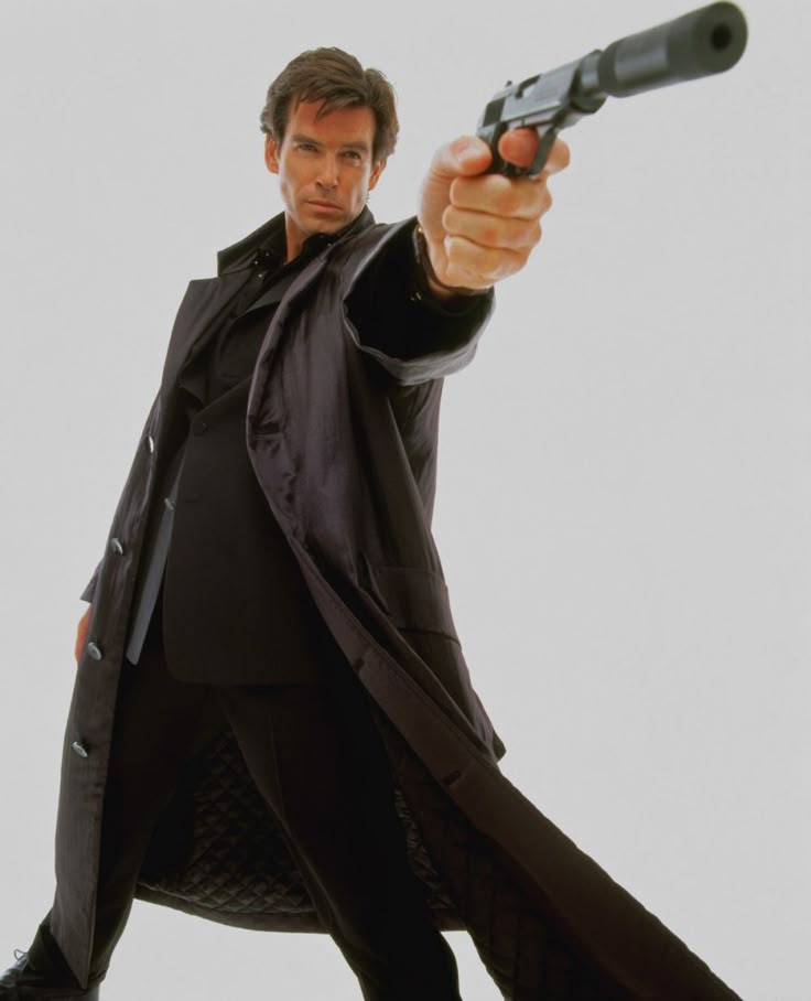 the image features pierce brosnan as james bond in the film goldeneye. he is wearing a black suit and coat, and he's holding a handgun with a silencer. he is pointing the gun directly at the viewer. he is posing in a studio setting.