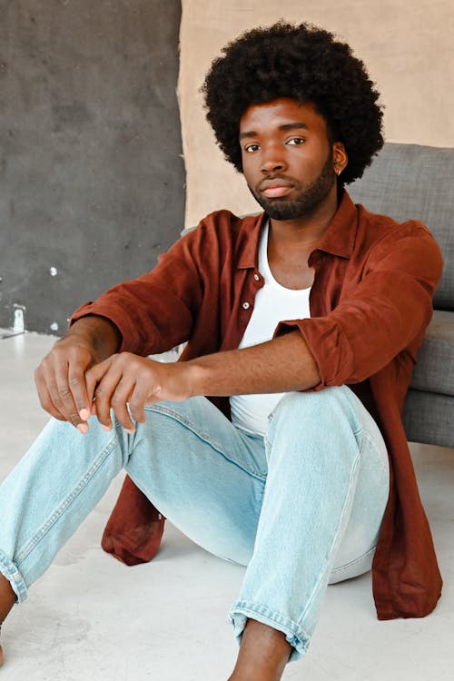 a handsome man with an afro is sitting on the floor in a relaxed pose, looking directly at the camera. he's wearing a white tshirt, a brown unbuttoned shirt, and light blue jeans. the setting appears to be a studio, with a simple backdrop of gray and beige. he has a beard and is barefoot, adding to the casual and confident vibe.