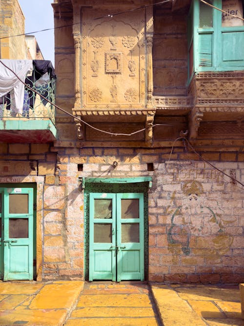 the image shows a closeup of a building with architectural details. the building features a stone wall with two turquoise doors sidebyside. there are windows with turquoise frames as well. a balcony with hanging laundry is visible on the left side of the image. the building has carved decorations on its surface. the architecture style and color scheme suggest that the building is located in india, possibly in jaisalmer.