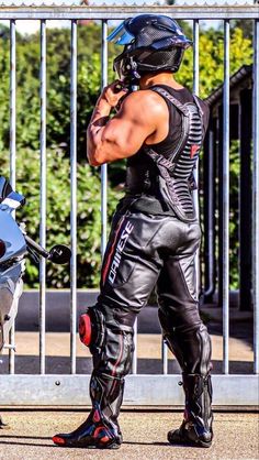 a muscular man wearing a black motorcycle helmet and full protective gear, including a leather vest, pants, and boots, stands beside his motorcycle. he is adjusting his helmet while looking away from the camera. the background features a metal gate and foliage, suggesting an urban setting. the sunny lighting highlights the man's physique and the details of his gear.