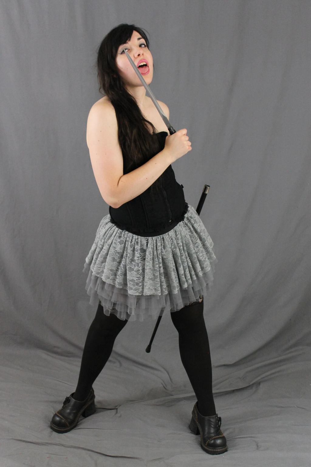 a fulllength shot of a young woman in a black corset and a layered grey lace tutu skirt.  she is wearing black tights and chunky black shoes. she holds a long, thin, silver object, possibly a sword or cane, playfully near her mouth with her mouth slightly open.  the background is a simple grey backdrop. the overall style is a blend of gothic and whimsical.