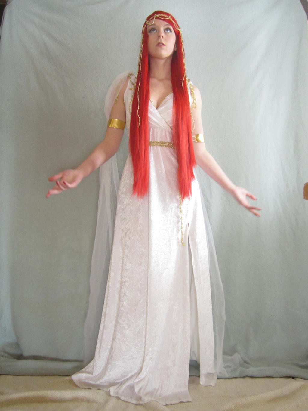 a fulllength shot features a woman dressed in a white gown, complete with a golden headpiece and armbands. her hair is styled in straight, fiery red tresses that contrast sharply with the white of her attire. the woman stands with her arms outstretched, palms facing upward, suggesting a gesture of offering or supplication.