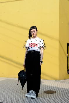 a woman in a stylish polka dot outfit stands against a yellow wall. she's wearing a white top with a black polka dot pattern, and her black pants  have a similar design. she's accessorizing with a black handbag and grey sneakers. she has a cool, casual style, and her pose is relaxed and confident. the bright yellow wall behind her creates a strong contrast, drawing attention to her edgy look.