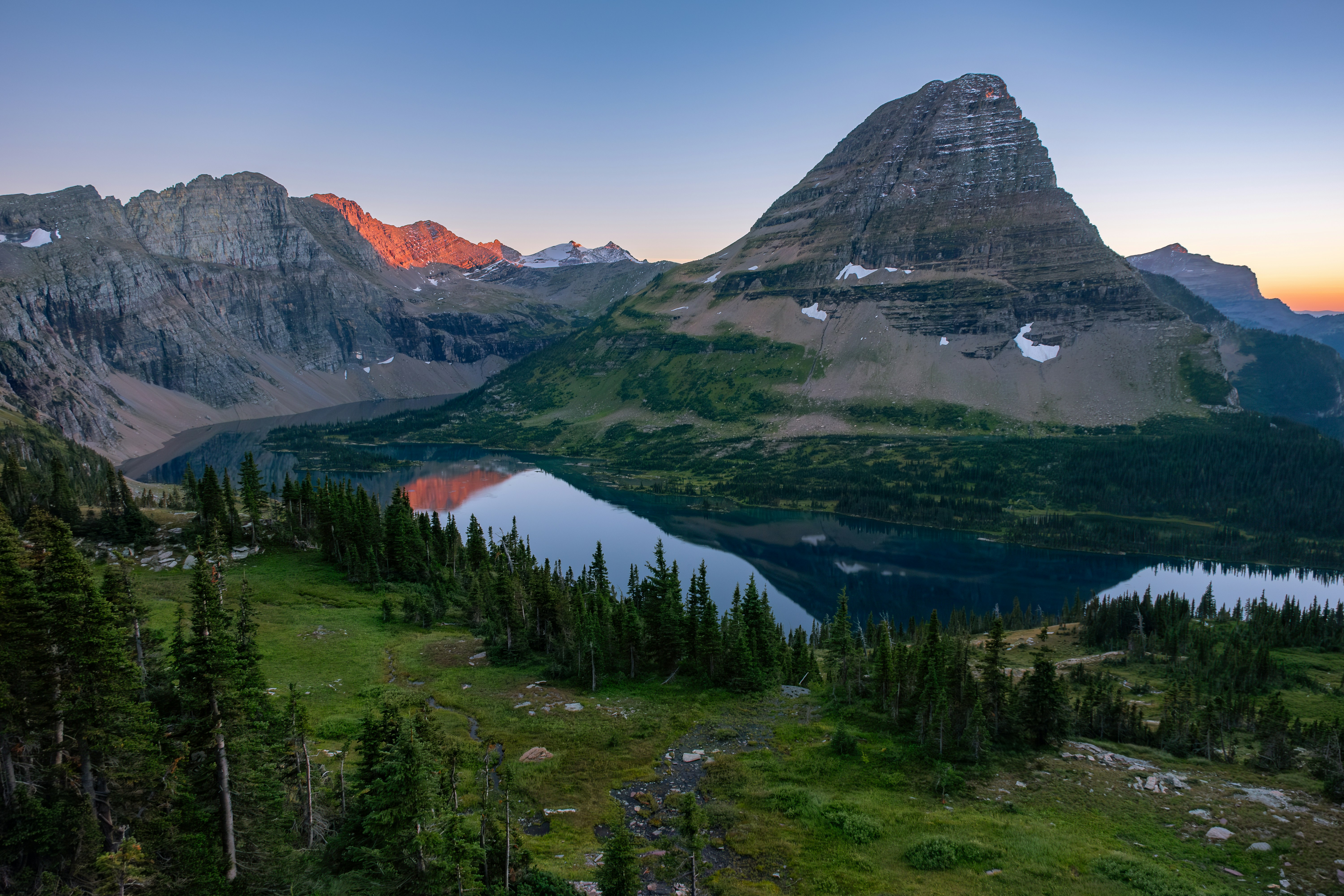 the image captures a breathtaking mountain landscape at sunrise. a serene alpine lake reflects the surrounding peaks, with the majestic mountain known as bearhat mountain prominently featured in the center. lush green forests cover the slopes, and patches of snow linger on the higher elevations. the sky displays a soft blend of blue and orange hues, casting a warm glow over the scene.