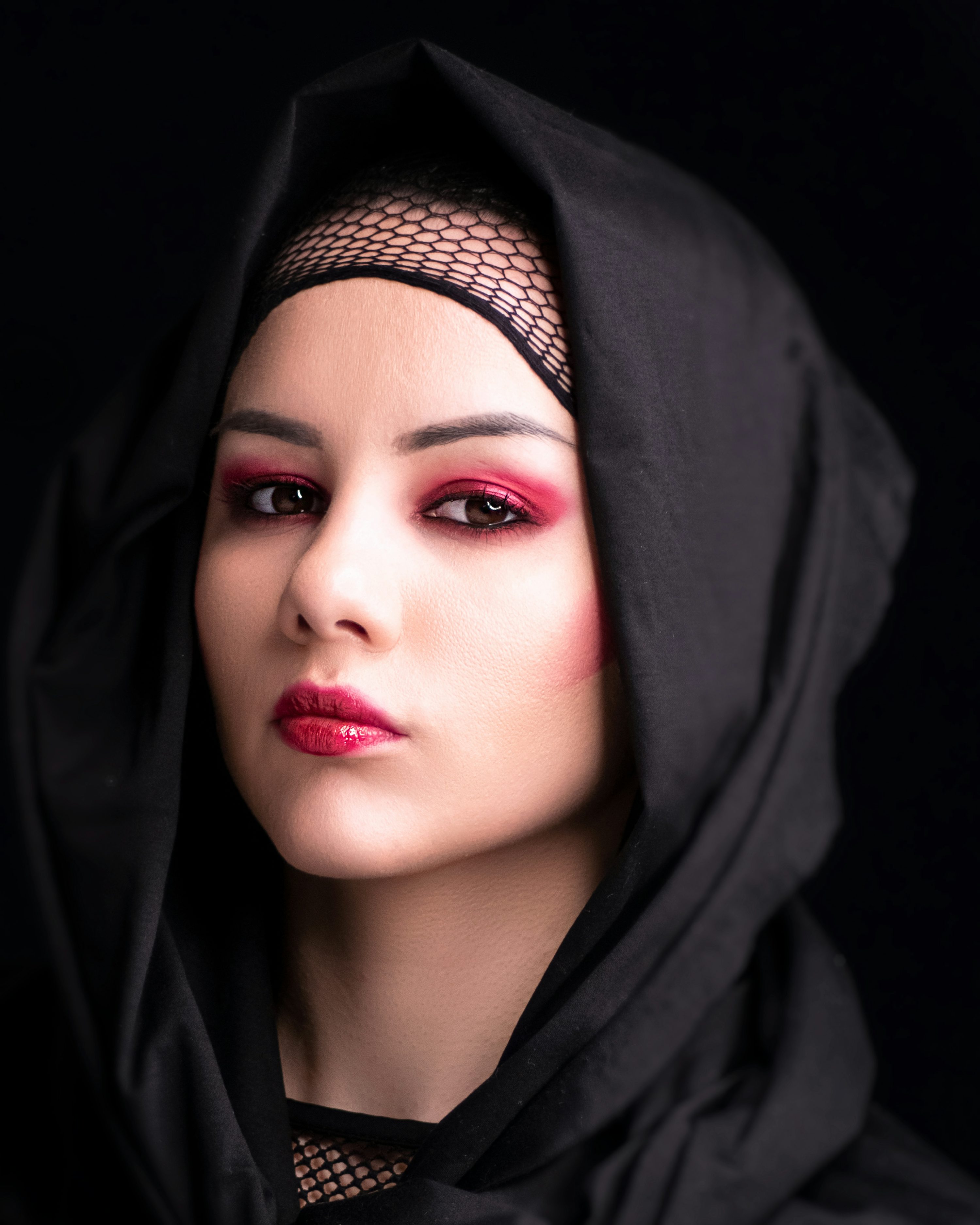 the image is a closeup portrait of a young woman. she is wearing a black hijab or headscarf that is draped around her head and covers most of her neck and shoulders. underneath, she has on a black fishnet top. her makeup is dramatic, featuring red lipstick and pink eyeshadow, making her eyes stand out against the dark background. the lighting is focused on her face, creating shadows and highlights that enhance her features. the overall mood is one of mystery and elegance. the woman's expression is serious and her gaze is direct, engaging the viewer.
