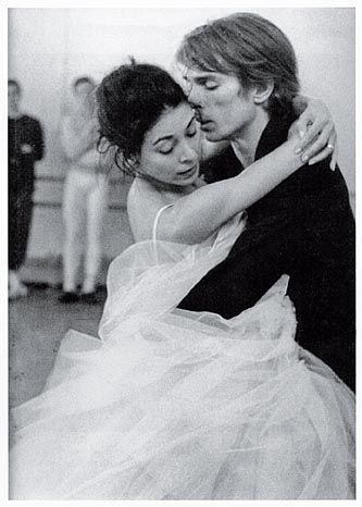 a black and white image of a couple dancing. the woman is wearing a beautiful white dress and the man is wearing a dark suit. they are embracing and seem to be deeply in love. the background is out of focus and only a few figures can be seen.