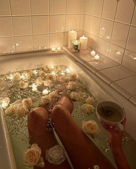 the image shows a person relaxing in a bathtub filled with water, white rose petals, and small white flowers. the tub is lit by numerous candles placed around the edge, creating a warm and calming atmosphere. the person is holding a cup of tea in their hand.