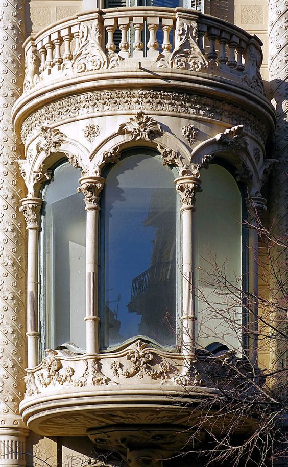 this is a closeup view of an ornate baroque balcony, featuring intricate carvings and a curved glass window.  the balcony's white stone facade stands out against the backdrop of a clear blue sky. the reflection in the window suggests an urban environment with another building visible. a bare tree branch extends into the frame from the right side, adding a touch of nature to the scene.