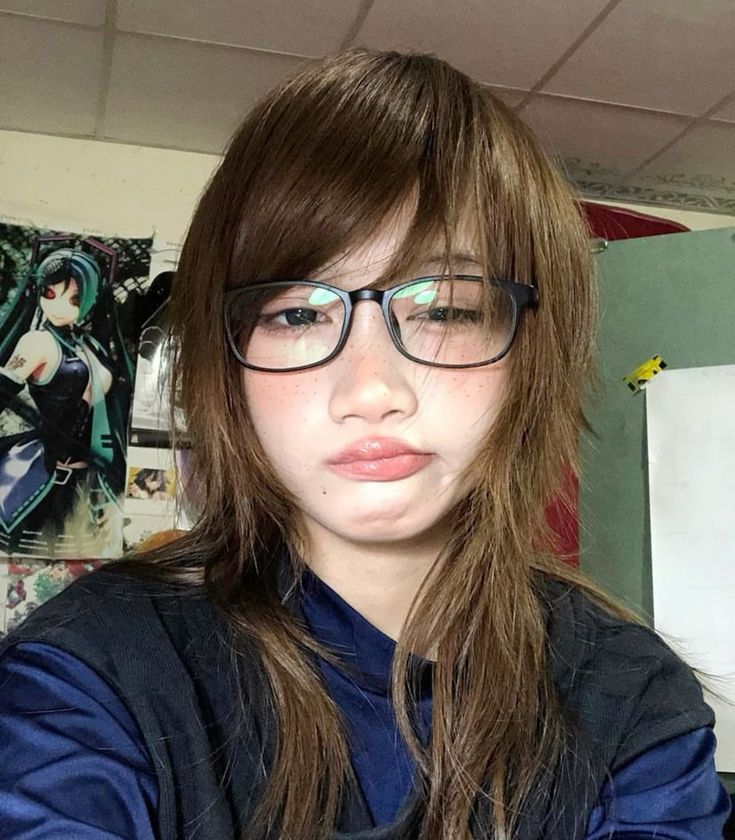 a young woman with long brown hair and glasses is taking a selfie. she has a bored expression on her face and is wearing a blue shirt. there is an anime poster on the wall behind her.