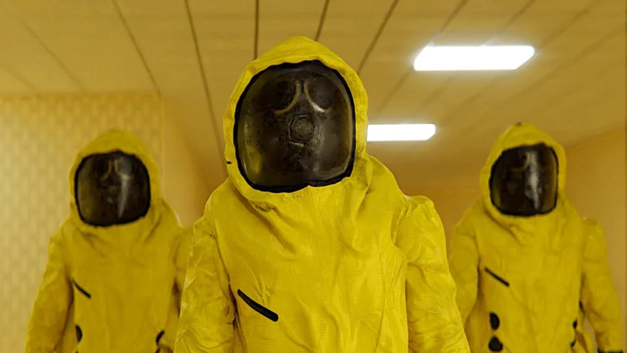 three people wearing yellow hazmat suits with respirators walk down a hallway, facing the camera. the suits are bright yellow and the overhead lights are very bright. the background appears to be a pale yellow.