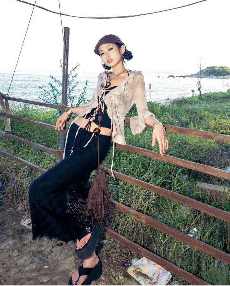 the image shows a young asian woman, styled in 90sinspired grunge/boho fashion. she's wearing a beige laceup top, a black lacedetailed maxi skirt, platform sandals, and a brown hat. she has long, dark hair styled in a halfupdo.  the model is leaning against a rustic metal fence, with a coastal background of a beach and calm ocean visible in the distance. the overall mood of the image is relaxed and casual, showcasing the model's stylish attire. the setting appears to be a serene outdoor location on a sunny day.