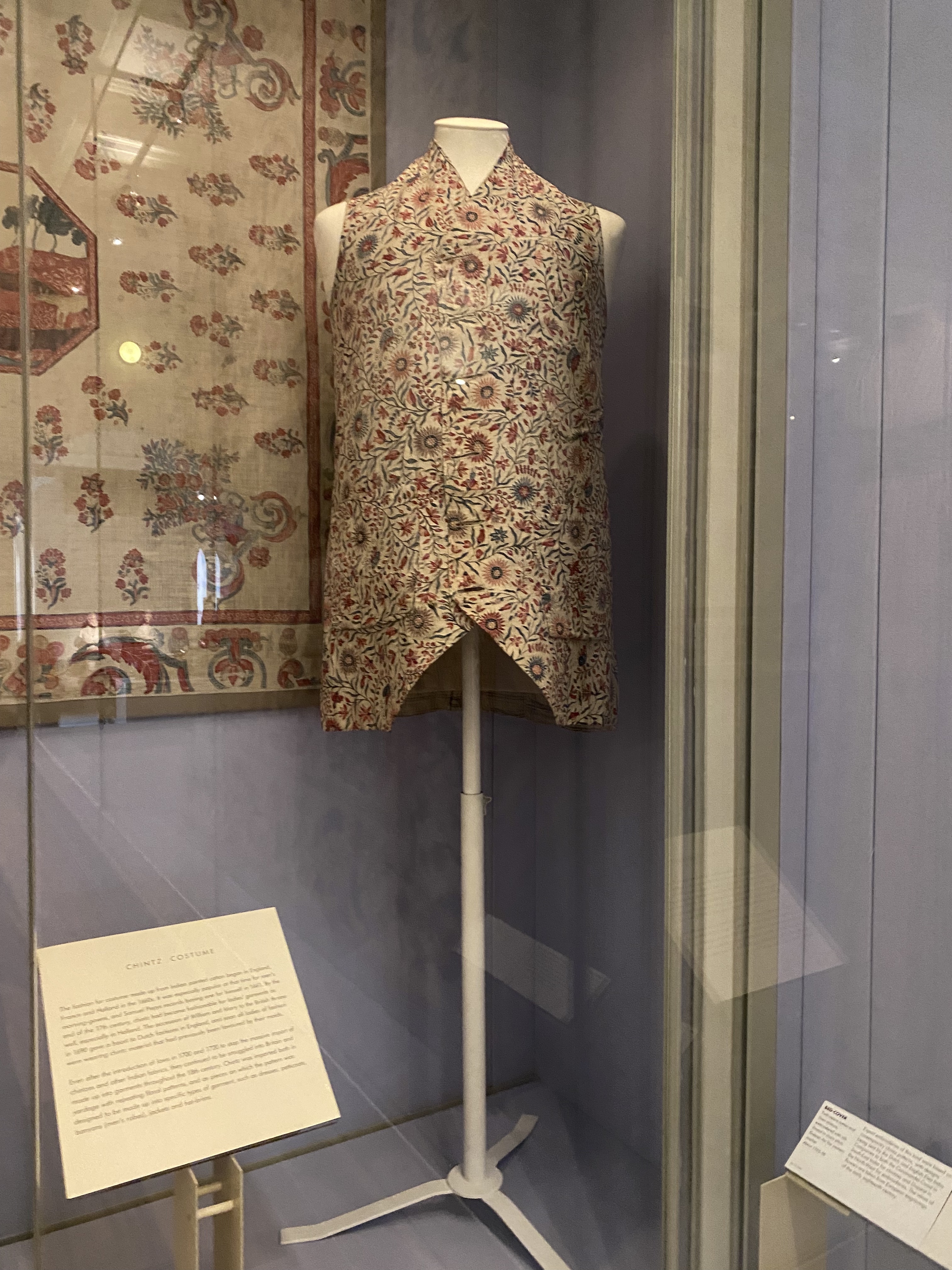 the image shows a floral patterned vest on a mannequin in a museum display case. next to the vest is a piece of textile also with a floral pattern. the overall lighting is bright, and the background is a pale blue color. a sign sits in front of the display.