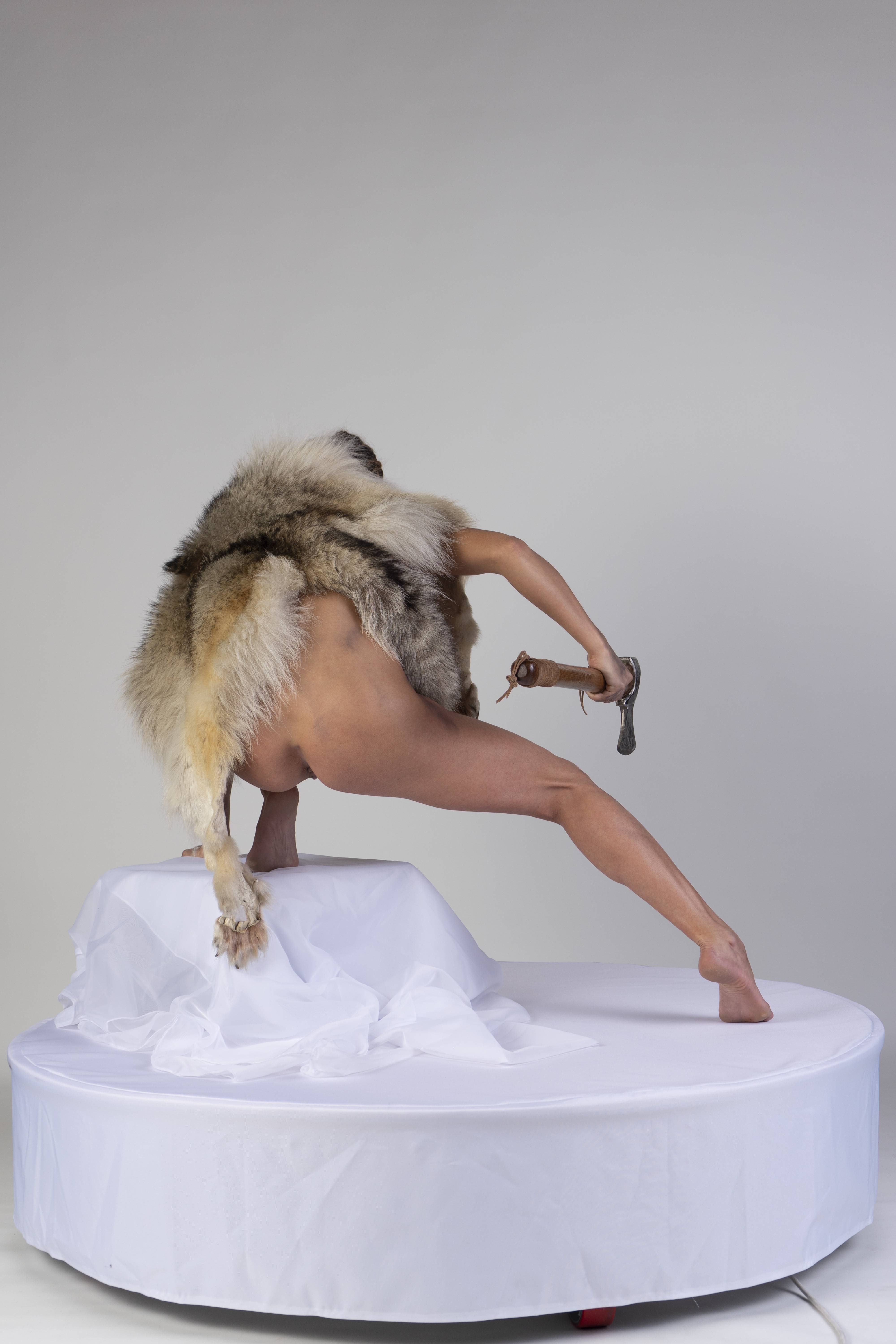 a nude woman wearing a wolf fur pelt stands on a white stage, holding an axe. the background is a plain white studio.