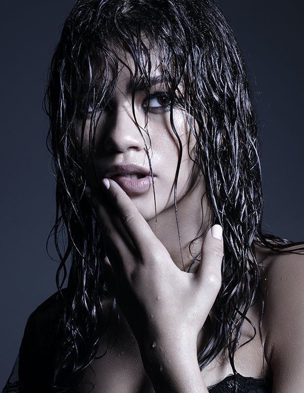 the image is a closeup, moody portrait of a young woman with long, wet, dark hair. her hair is styled in a way that frames her face, with strands hanging down. water droplets are visible on her skin and hair.  she has a pensive expression and is touching her face with her hand. the lighting is dramatic, creating shadows and highlighting her features. the background is dark and unfocused, drawing attention to the woman. the overall mood is sensual and mysterious, with a touch of elegance. the woman appears to be wearing minimal makeup, enhancing her natural beauty. she is mostly bare shouldered.