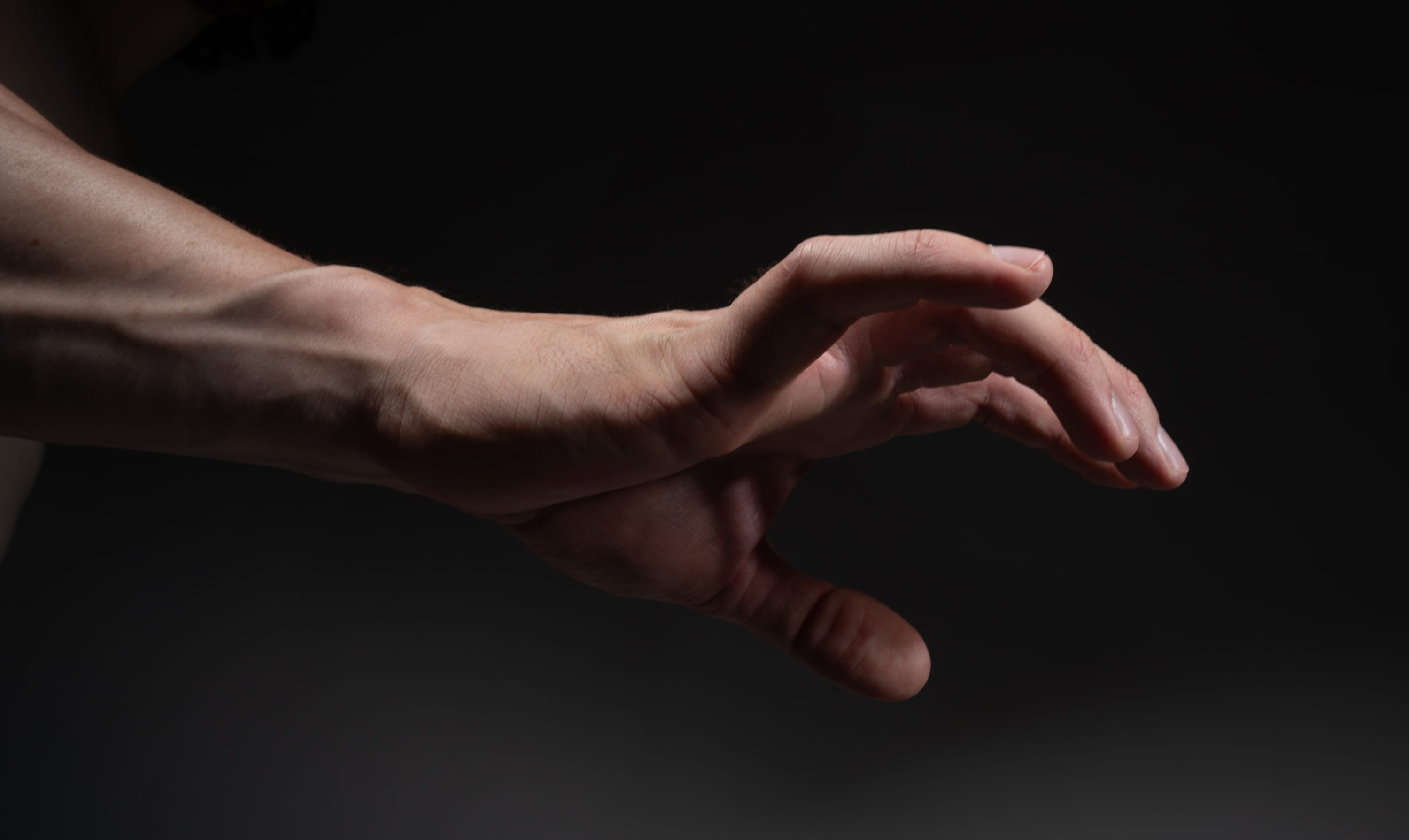 a closeup shot of a man's muscular arm extended forward with an open hand, as if reaching out to grasp something. the studio lighting highlights the contours and details of the hand and arm, emphasizing the veins and muscles. the dark background creates a sense of drama and intensity.
