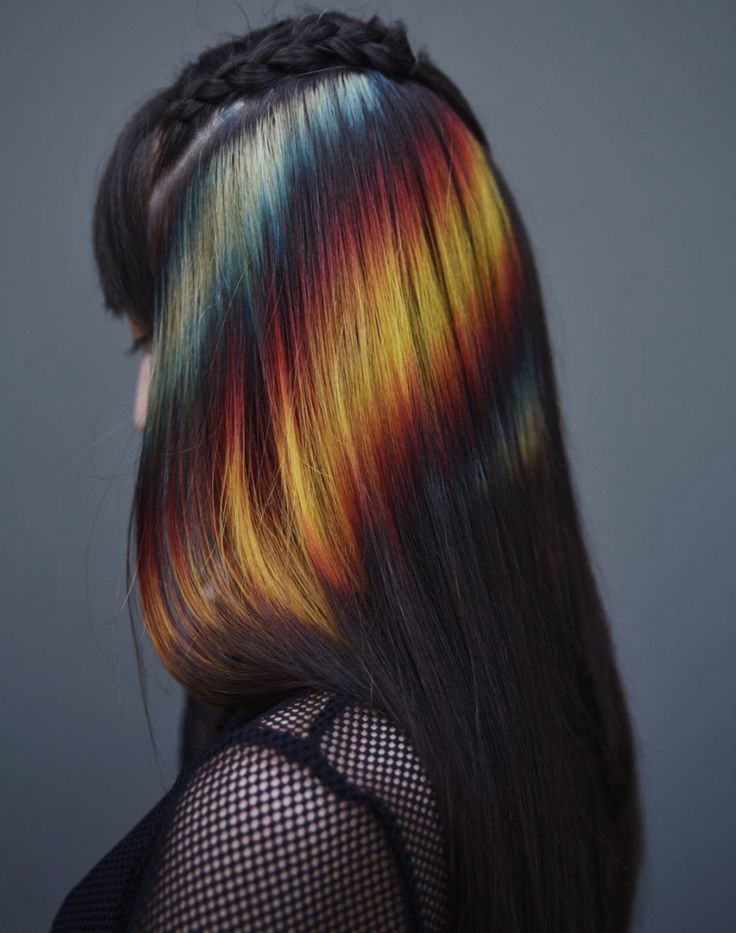 a woman with long black hair has colorful streaks of yellow, red, and blue in her hair. her hair is braided on top and she is wearing a black mesh shirt. she is looking away from the camera and the side of her hair is showing.