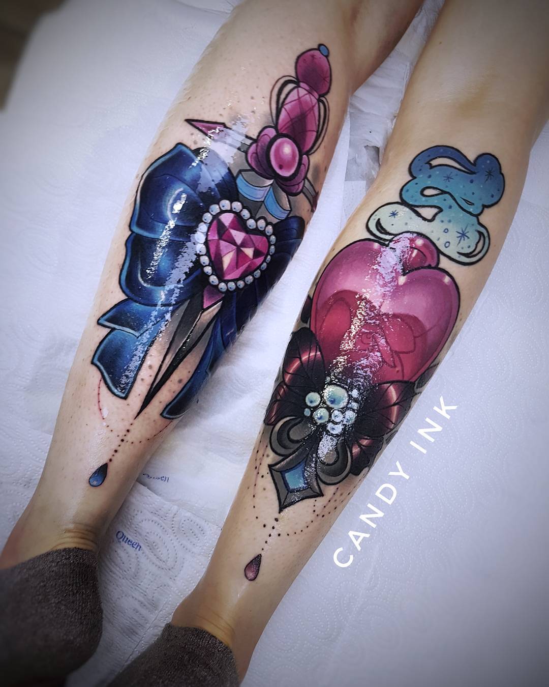 a closeup image featuring two neotraditional tattoos on a person's legs. the first tattoo depicts a jeweled dagger with a blue bow. the second tattoo portrays a pink heart adorned with jewels and a cross. the tattoos are vibrant and detailed, showcasing a blend of colors and intricate linework. the image captures the artistry and visual appeal of these body art pieces.