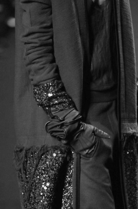 a black and white photo of a woman wearing a stylish long black coat. the woman is standing and the image is taken at a studio.  she is also wearing black leather gloves with sequins on them. the outfit is very elegant.