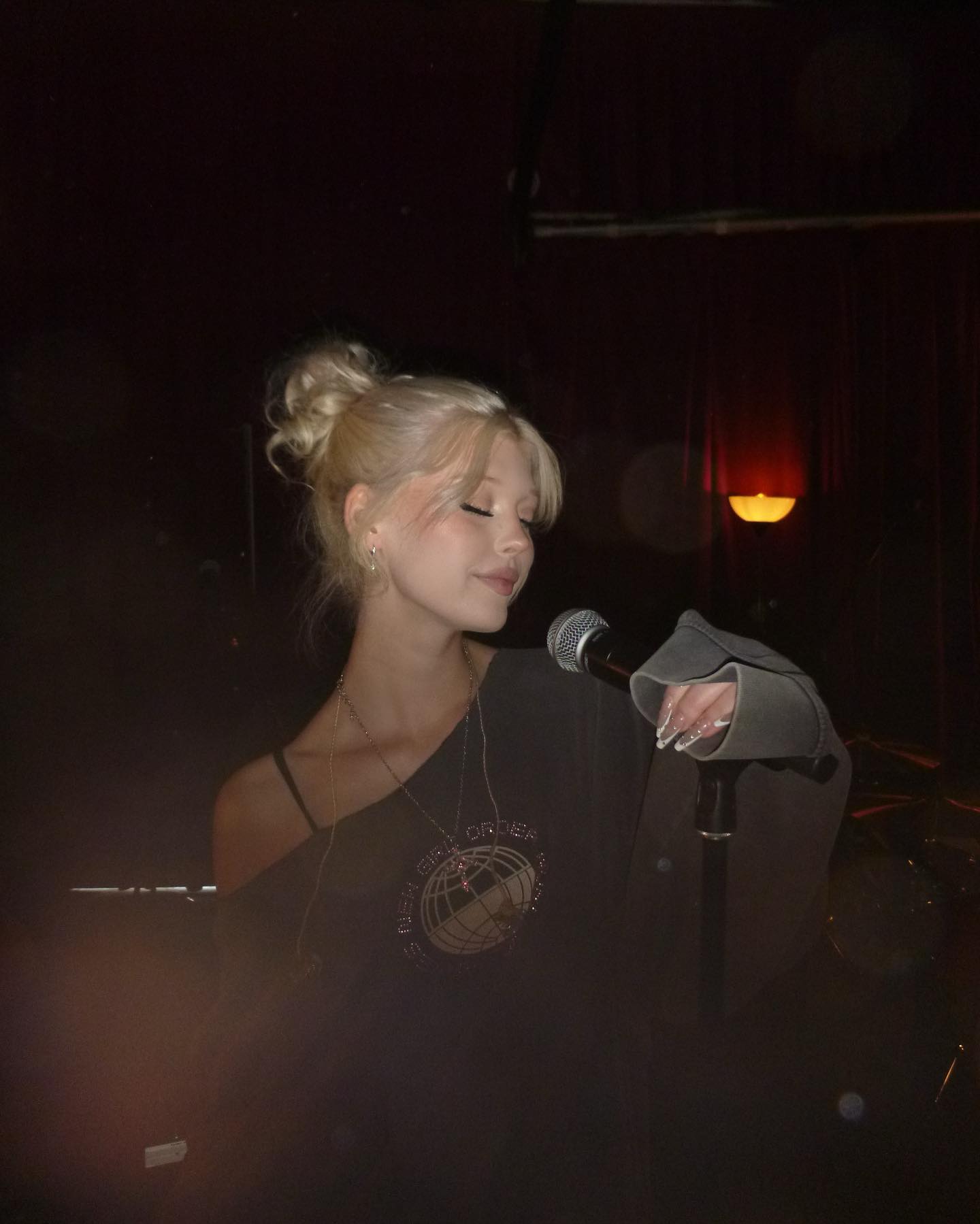 a woman with blonde hair styled in a bun is standing on a stage, singing into a microphone. she's wearing an offshoulder top with a "new girl order" design and several pieces of jewelry, including a necklace. the background is dark and blurred, with some stage lighting visible. her eyes are closed.