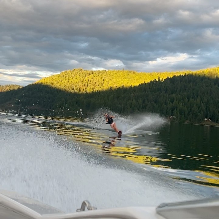 a girl water skis across a lake at sunset. the lake is surrounded by green mountains, and the sky is a mix of clouds and clear blue. the girl is wearing a black top and white bottoms and is enjoying the sport. the wake from her skis is visible in the water, and there's a hint of a boat in the lower left corner of the image. the sunlight reflects beautifully on the water.