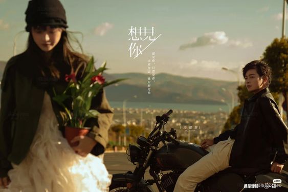 a young couple is standing beside a black vintage motorcycle on a road with mountains and a lake in the distance. the woman is holding a potted plant and the man is sitting on the motorcycle. the sun is setting in the background, casting a warm glow on the scene. the image has a vintage feel with a muted color palette.