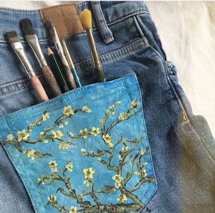 a pair of blue denim jeans lies flat with the back pocket facing the camera.  an assortment of paintbrushes and pencils are tucked into the pocket.  the pocket is adorned with a handpainted reproduction of vincent van gogh's "almond blossom" painting.