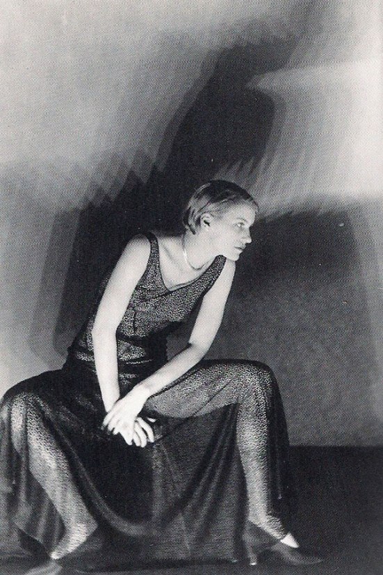 a black and white photograph of a woman in a long, sheer black dress. she is sitting with her knees bent and her hands resting on her lap. the dress is formfitting, and the fabric appears to be a delicate mesh or net. the woman's hair is styled in a short, bobbed cut, typical of the 1920s or 1930s. the background is a plain, dark wall, with the shadows of the woman cast on the wall behind her, creating a dramatic effect. the photograph is beautifully lit, highlighting the texture of the dress and the woman's features. the overall mood of the image is mysterious, elegant, and sensual.
