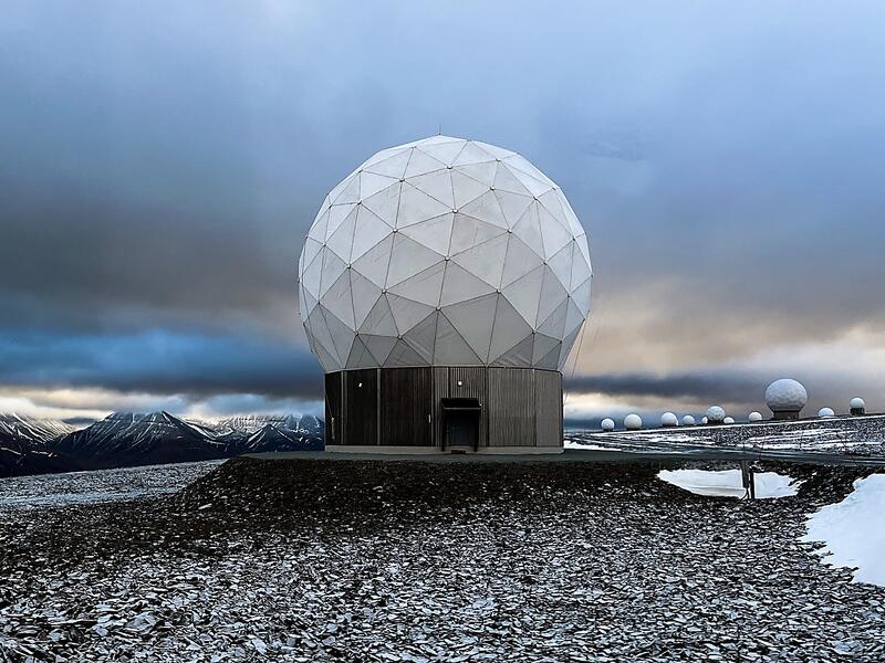 a large white radar dome stands on a snowy landscape in front of a mountain range. the dome is a geodesic structure with a wooden base. there are several smaller radar domes in the background, arranged in a line. the sky is cloudy and blue.