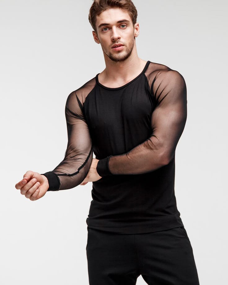 a man is standing in a studio with a white background. he is wearing a black longsleeved shirt with mesh sleeves and black pants. the shirt is a slim fit. the man is looking at the camera.