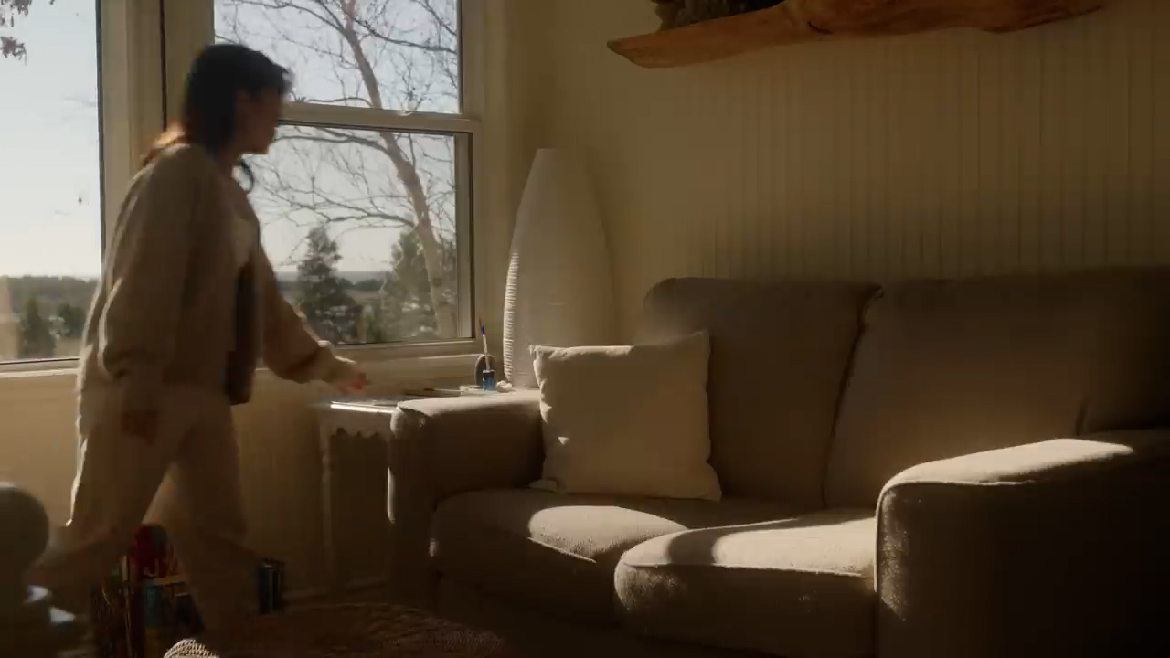 a woman walks past a sundrenched couch in a cozy living room. the couch is a light beige color, and the room is decorated in neutral tones. sunlight streams in through the window behind the couch, illuminating the woman's silhouette.  a small side table sits in front of the window.  the overall atmosphere is warm and inviting.