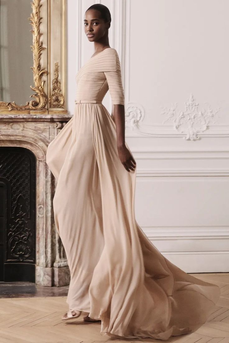 a woman with short black hair is wearing a long beige gown. the dress has a slight vneck and is sleeveless. the gown has a flowing design and the bottom of the dress is trailing the floor. the woman is posing in a studio with a white background and the wood floor is visible in the bottom of the frame.