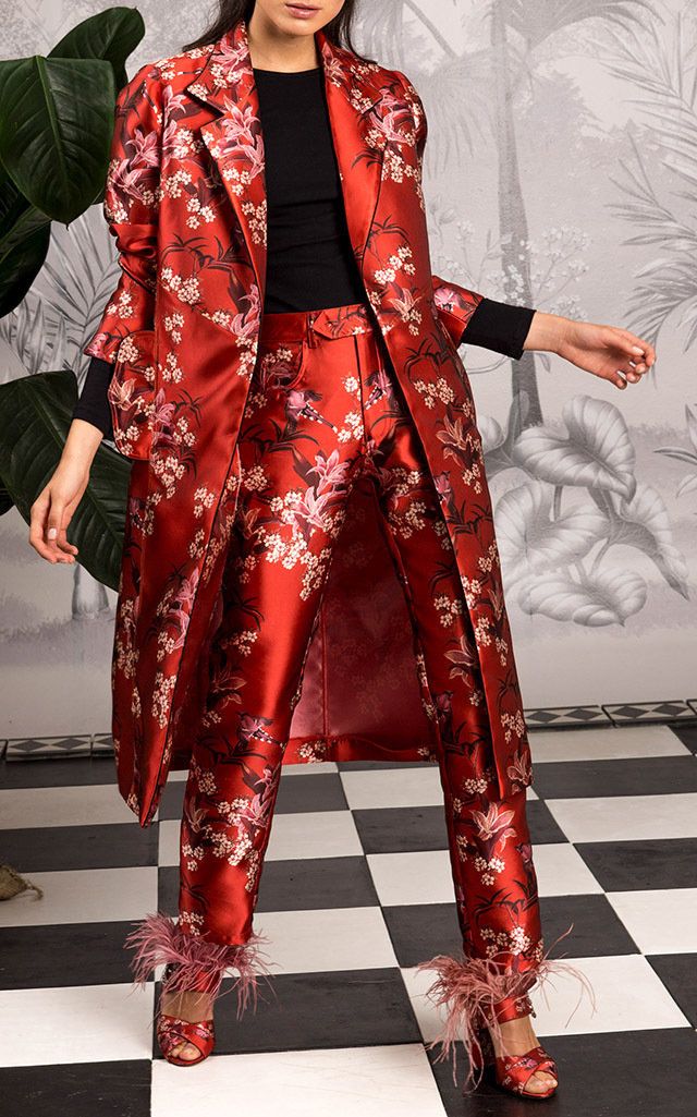 a fulllength shot of a woman showcasing a stunning red floral pantsuit. the outfit comprises a long, loosefitting coat and matching trousers, both adorned with an intricate cherry blossomlike print on a rich red satin or silk material.  the model wears black top underneath and elegant highheeled shoes embellished with feathers.  she is posed on a black and white checkered floor, with lush green plants in the background. the overall aesthetic is sophisticated and luxurious.