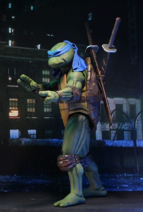 a closeup of a teenage mutant ninja turtles action figure. the figure is of michelangelo, and he is posed in a fighting stance. he is wearing his signature blue bandana and holding his two katanas. he is also wearing his turtle shell, and his green skin is visible. he is standing on a dark background.