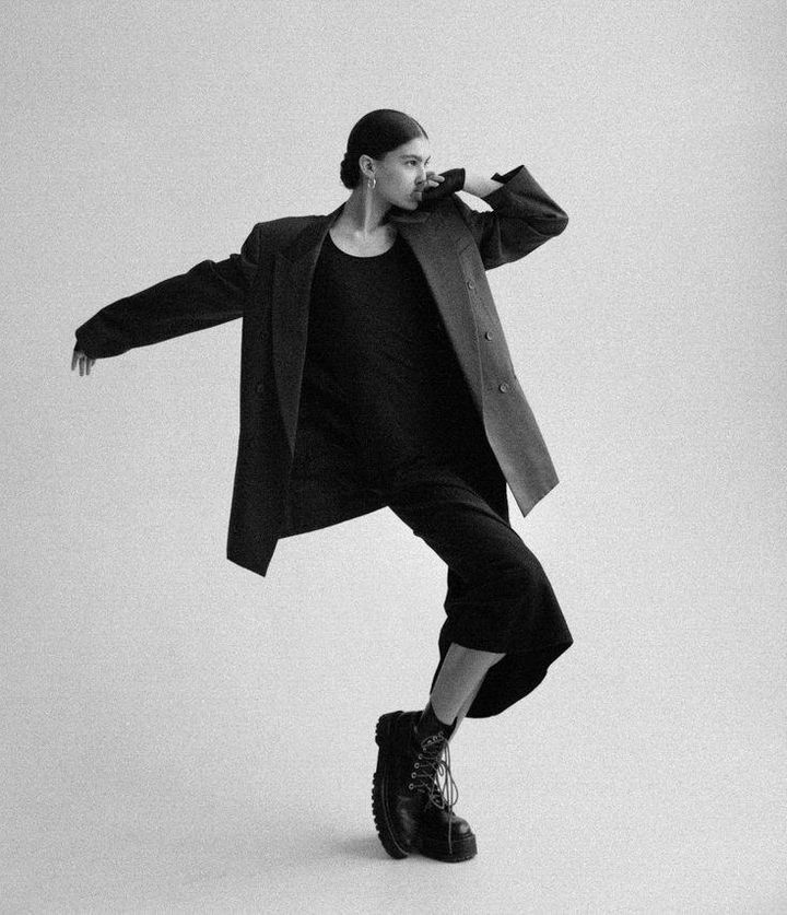 a black and white full shot of a stylish woman. she is wearing a black blazer, black fitted jumpsuit and black combat boots. the woman is posing dynamically, one leg bent, her arm extended to the side. the backdrop is a simple, lightgrey studio background.  the overall feel is modern, edgy, and minimalist.
