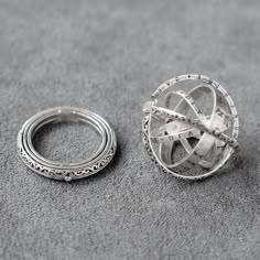 a close up of two silver rings. the ring on the left is a simple band with an intricate, engraved design. the ring on the right is a celestial sphere with a detailed, multilayered design. both rings are on a grey background.