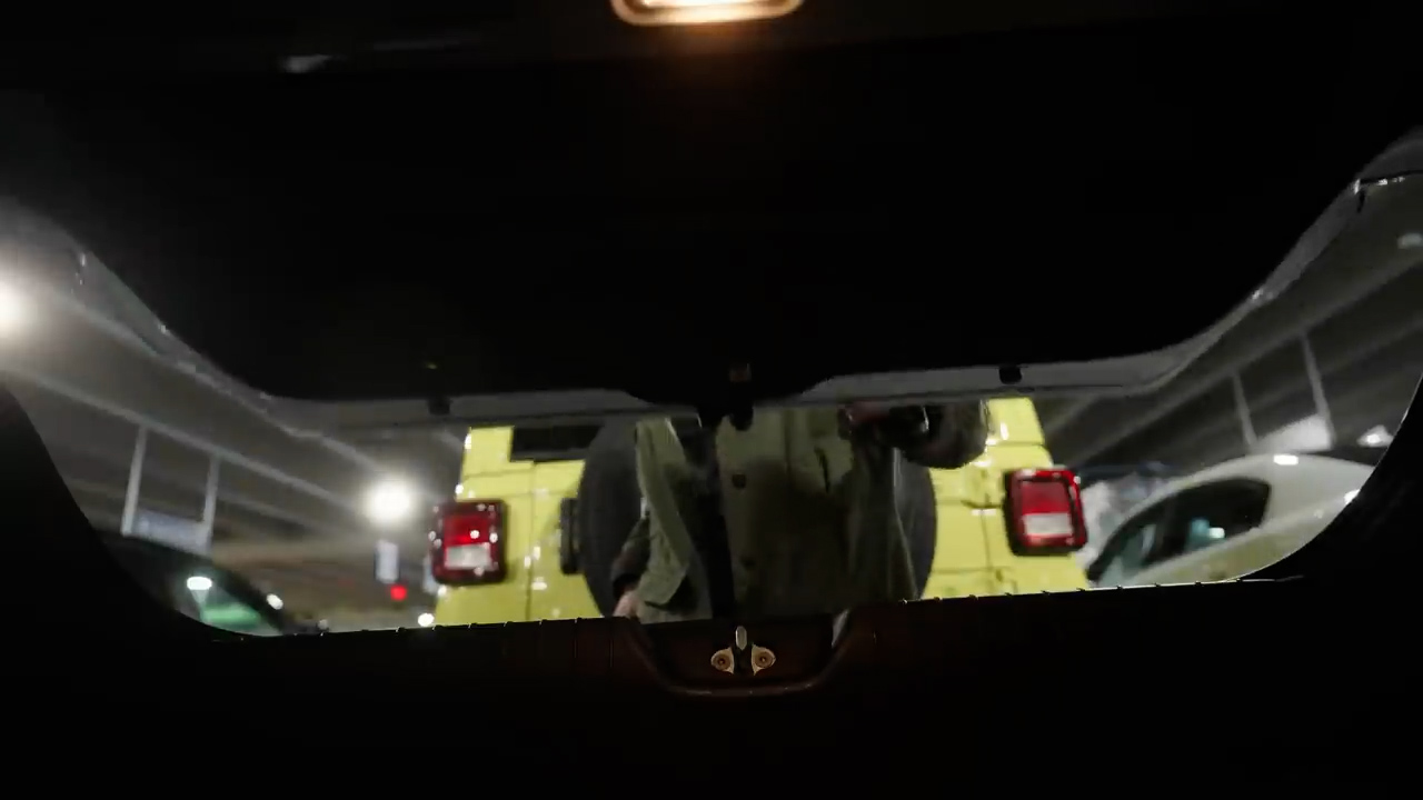 the image shows the view from inside a car trunk that is open.  the trunk is dark and the parking garage in the background is dimly lit.  a person is standing in the background and is partially obscured by the trunk lid.  the person is holding a red object in their right hand.  a yellow jeep wrangler is visible in the background to the left of the person.