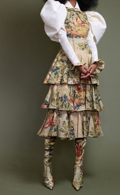 a full shot of a fashion model wearing a unique and elaborate outfit. the dress is a tiered, beige skirt with a multicolored floral tapestry print. the top half consists of a white shirt with dramatic puff sleeves. the model is wearing matching high boots with the same floral tapestry print. the background is a muted olive green. the overall style is vintageinspired, with a modern twist. the image appears to be a professional photoshoot, possibly for a fashion magazine or runway show.