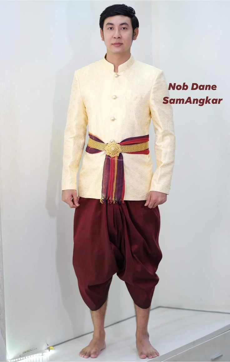 a man is standing wearing a traditional thai outfit for a formal occasion. he is wearing a white shirt with gold buttons and gold trim, maroon pants, a gold sash with a gold buckle and a red and gold fabric sash around his waist. he has a serious expression and is looking at the camera. the background is white.