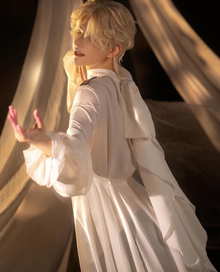 a blonde haired model with a white dress poses elegantly in a studio with soft lighting. the dress features intricate details and a flowing skirt. their expression is ethereal and dreamlike.