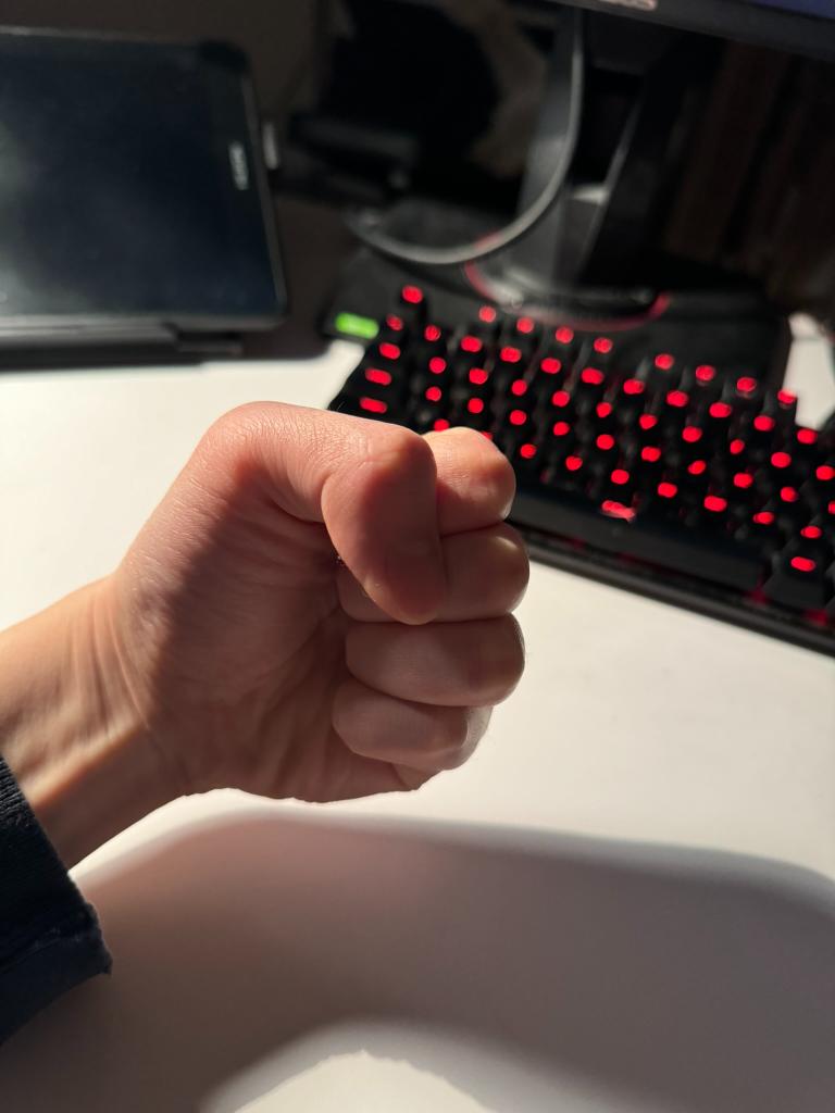 he image shows a person's hand clenched into a fist in front of a black mechanical keyboard with red backlit keys. the hand is resting on a white desk. the lighting suggests natural light coming from the left, casting a shadow of the fist onto the desk. the perspective is from the point of view of the person, as if they are looking down at their own fist. the overall impression is one of tension, possibly frustration or anger, perhaps related to using the computer or playing a game.