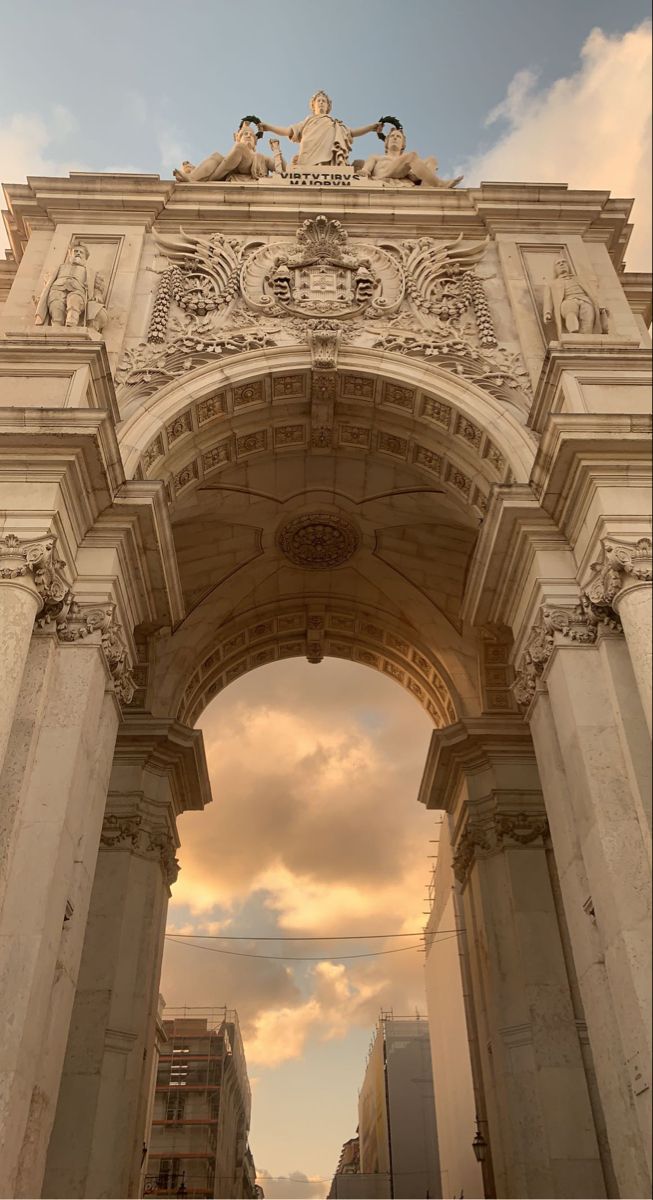 the image showcases a detailed view of the rua augusta arch in lisbon, portugal. the arch, a prime example of classical architecture, is captured during the golden hour, casting a warm, beigetoned light across its intricate carvings and statues. the sky above is a blend of soft clouds and a setting sun.  the arch's structure is emphasized, highlighting its columns, ornate detailing, and the sculptures atop it.  the surrounding cityscape is partially visible, adding a sense of context to the monument. the overall aesthetic is serene and subtly luxurious, leaning towards a pastel color palette.