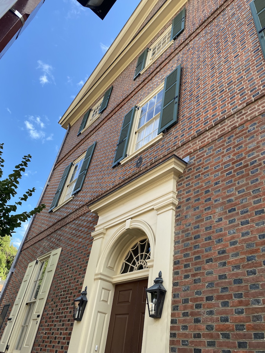 low angle view of a brick building with windows, shutters, and a front door. the building has a tan trim around the windows and door. there are lanterns flanking the door. the sky is visible in the background.