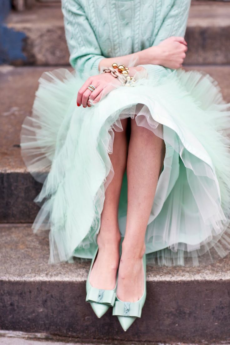 a woman is sitting on a set of stairs wearing a mint green cable knit sweater and a tulle skirt of the same color. she is wearing mint green bow flats and has several gold and pearl bracelets and rings on her hands. the overall aesthetic is elegant and feminine, with a spring/summer feel.